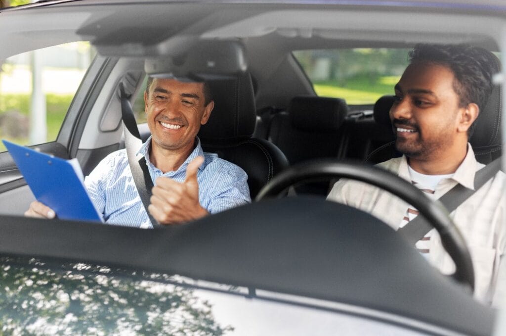 driver courses, exam and people concept - happy smiling indian man and driving school instructor with clipboard showing thumbs up in car