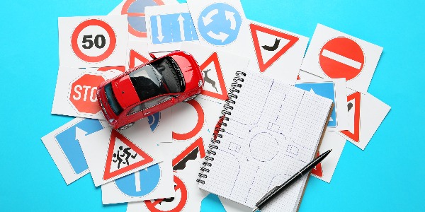 Many different road sign cards, notebook with sketch of roundabout and toy car on light blue background, flat lay. Driving school